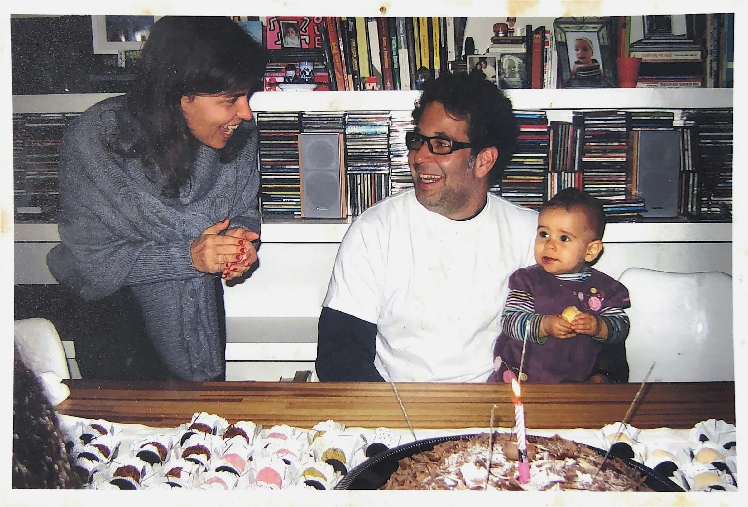 Familienfoto am Geburtstagstisch: Eine Frau und ein Mann lachen sich an, daneben sitzt ein Baby, vorne steht ein Kuchen mit Kerze.