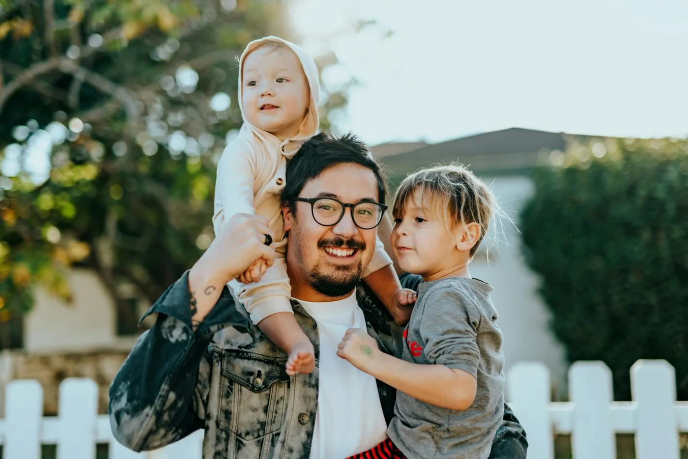 A father carrying a toddler on his shoulders, with a second child holding on to him.